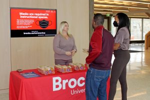 A woman stands behind a table draped in a Brock University table cloth. She is speaking with two people who are standing at the front of the table.