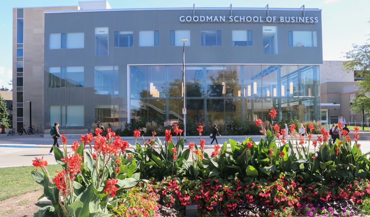 A three-storey grey building with large windows, hanging lights, and a sign at the top of the building that reads ‘Goodman School of Business.’