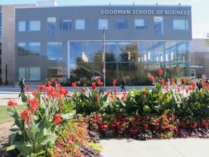 A three-storey grey building with large windows, hanging lights, and a sign at the top of the building that reads ‘Goodman School of Business.’