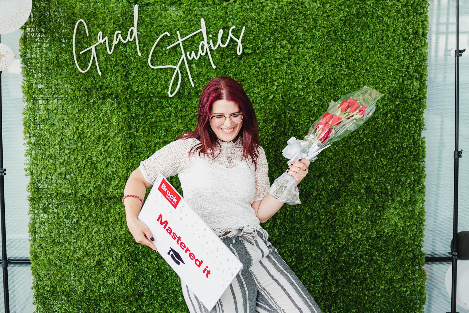 A Brock University graduate student celebrating while holding ‘Mastered it’ sign in front of faux green Graduate Studies photo backdrop during convocation celebration.