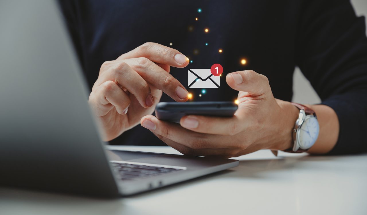 A person using both of their hands to check email on their smartphone. A digital illustration above the phone indicates the person has one unread email.