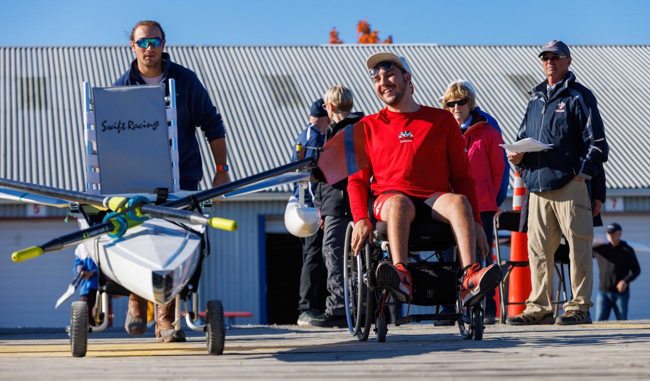 A man in a wheelchair travels along a dock with a modified racing boat with the help of teammates.