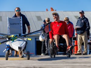 A man in a wheelchair travels along a dock with a modified racing boat with the help of teammates.