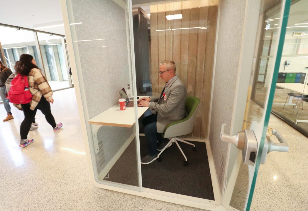 A man sits in a glassed-in meeting space working on a laptop.