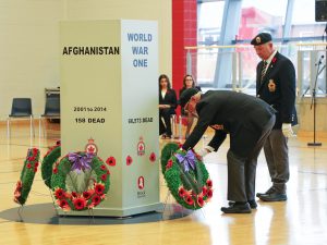 A man places a wreath at the base of a cenotaph while another man looks on. Both are in military uniforms.