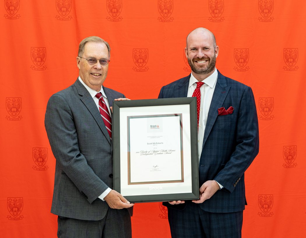 Applied Health Sciences Dean Peter Tiidus congratulates Scott McRoberts on his award accomplishment in front of a red wall.
