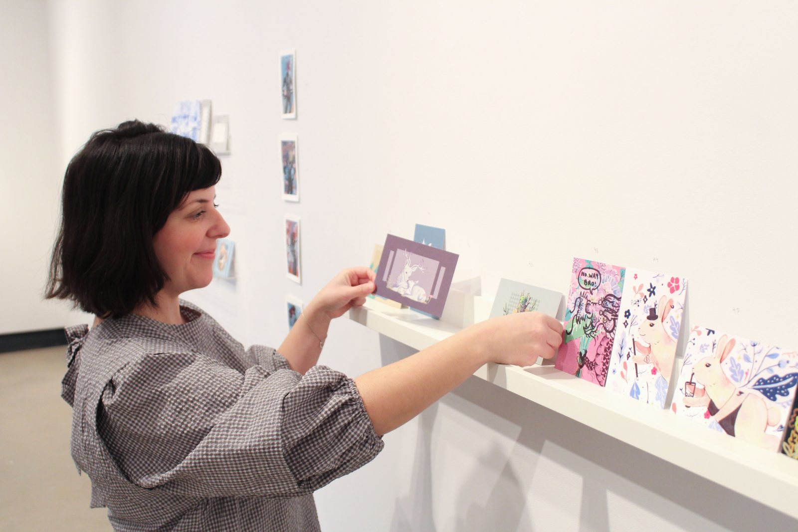 A female places a postcard on a shelf with other artistic creations in a gallery space.