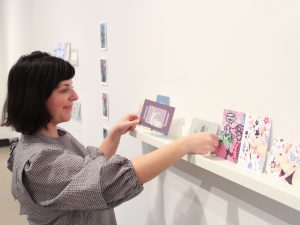 A female places a postcard on a shelf with other artistic creations in a gallery space.