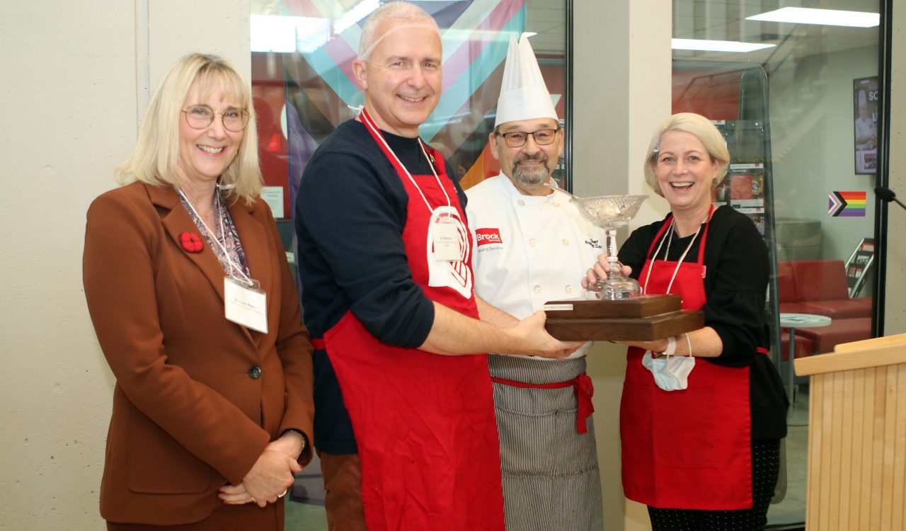 Four people stand next to each other holding a trophy that looks like a clear stemmed bowl with a ladle. One is wearing a chef's hat and whites, and two are wearing red aprons with the United Way logo on them.