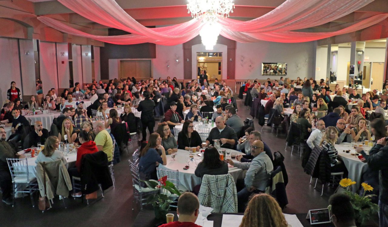 A large ballroom with a chandelier has approximately 20 round tables filled with 10 people at each table.