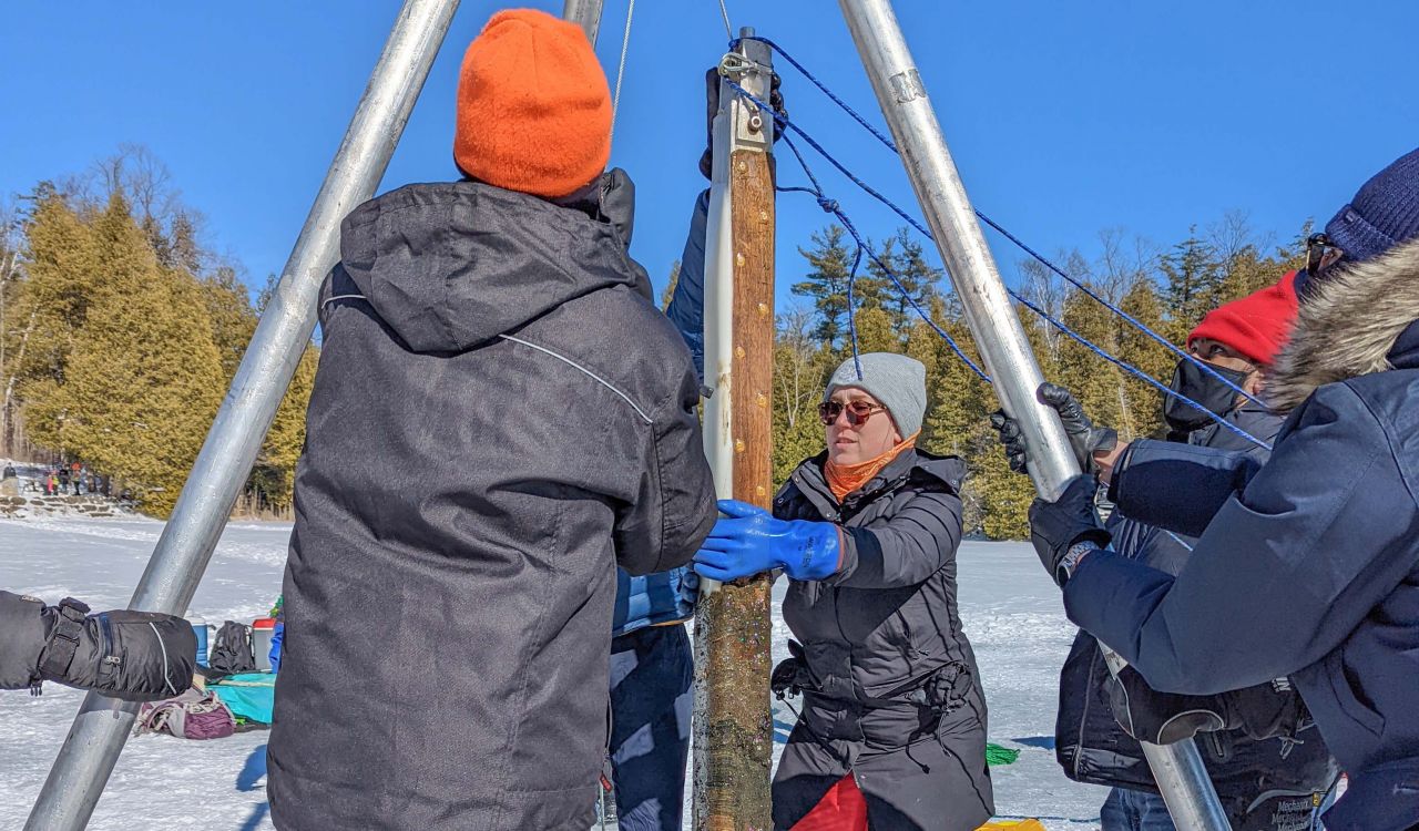 A group of people wearing blue winder jackets and gripping silver poles surround a brown tube that looks like a pole being pulled up from the snow-covered ice.