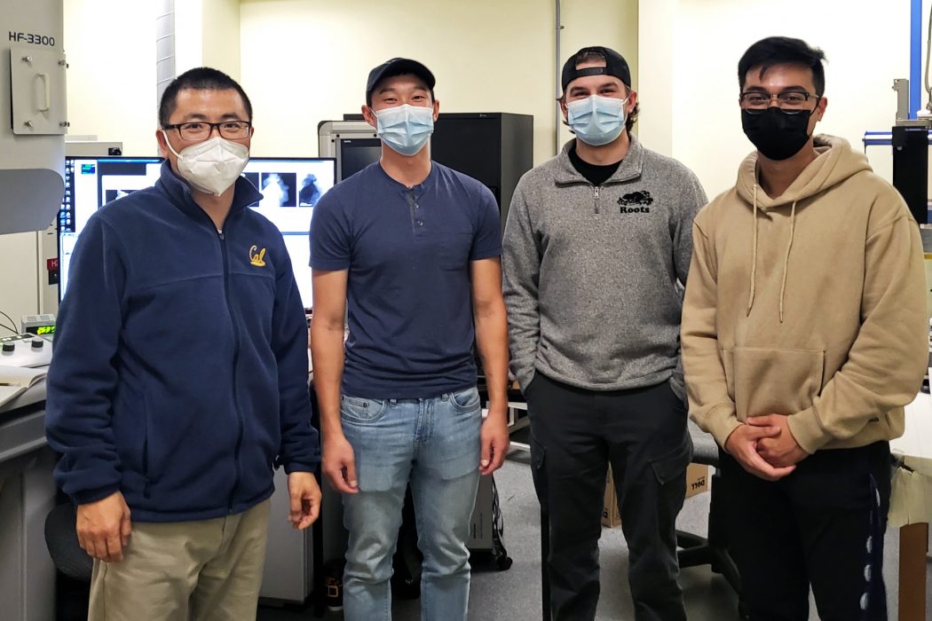 Three students and their professor pose for a photo while wearing medical masks. They stand in a line with computers and laboratory equipment in the background.