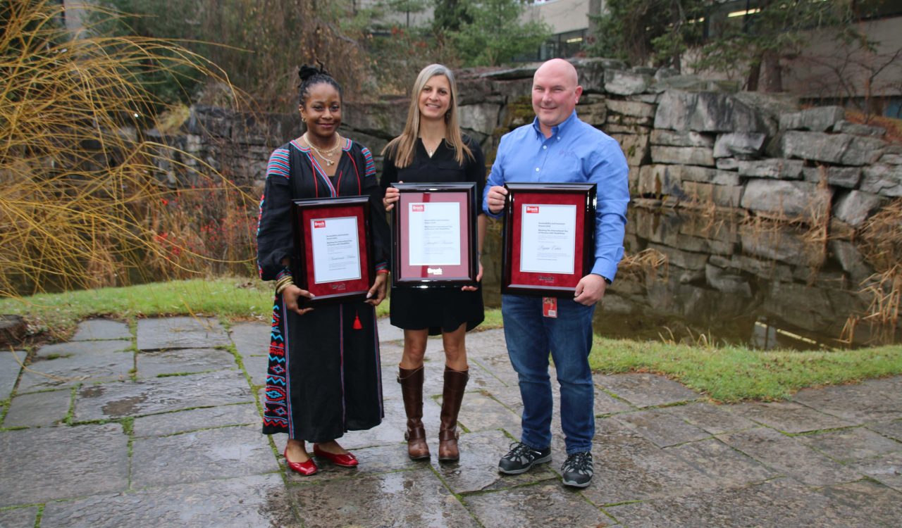 Two women and a man hold framed award certificates in front of an outdoor pond.