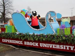 Brock University's mascot, Boomer the Badger, stands in a blue sleigh that sits atop a large holiday parade float. The sleigh is surrounded by oversized presents wrapped in colourful paper and bows.