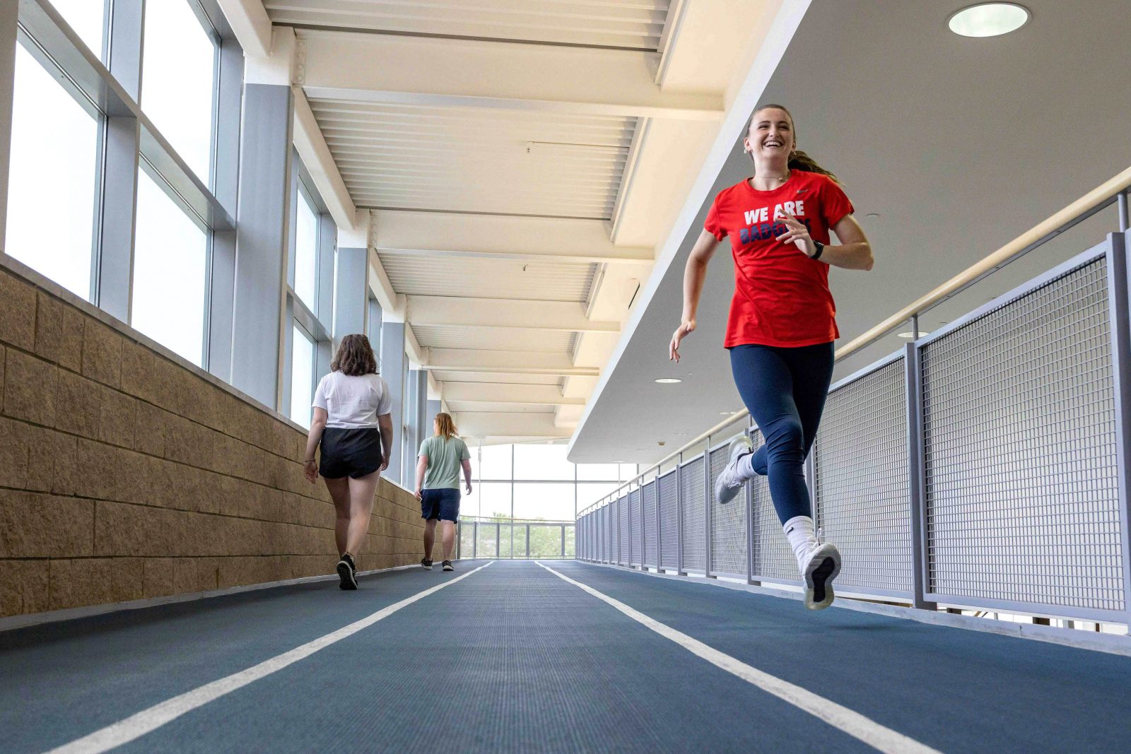A woman in fitness attire runs on an indoor track while others walk.
