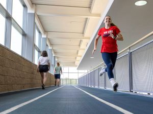 A woman in fitness attire runs on an indoor track while others walk.