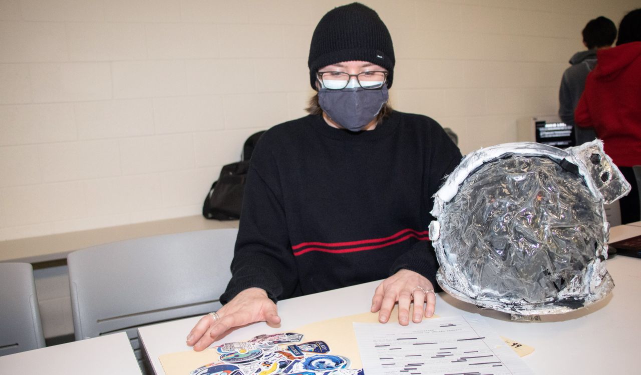Student sitting behind a mockup of a space helmet with documents and stickers on the table.