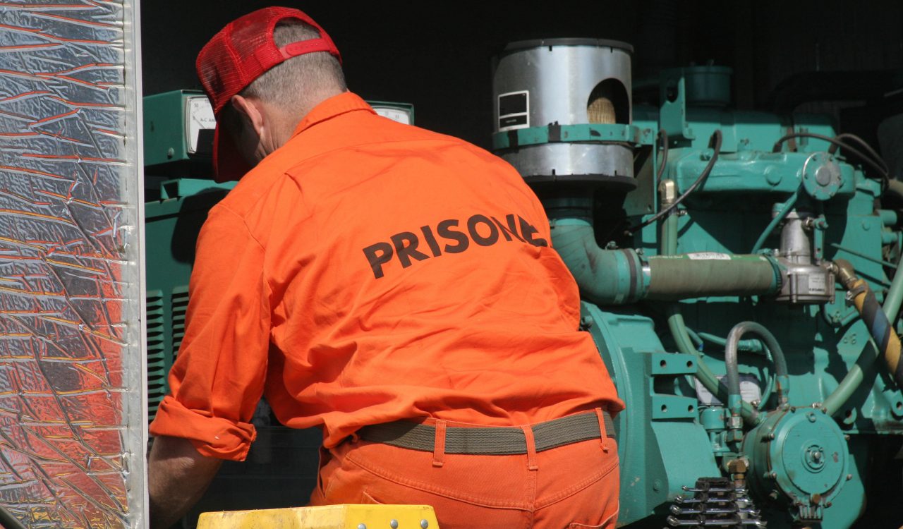 A man in an orange jumpsuit with “prisoner” printed across the back works on a large green machine.