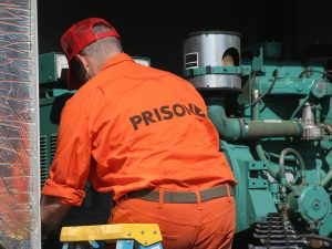 A man in an orange jumpsuit with “prisoner” printed across the back works on a large green machine.