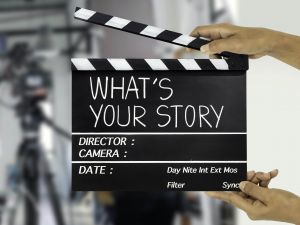 Director’s slate in foreground, saying “What’s Your Story?” with blurred background of cameras in a studio.