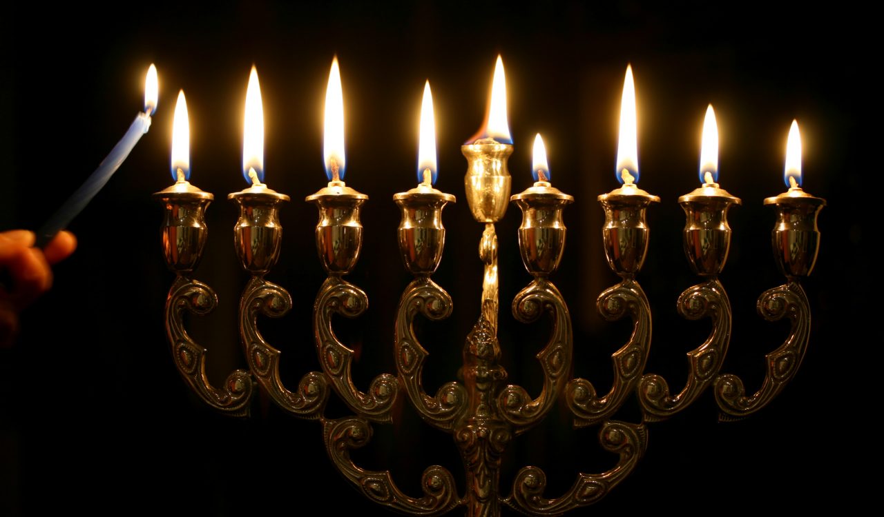 The nine wicks of a gold menorah burn brightly against a black backdrop.