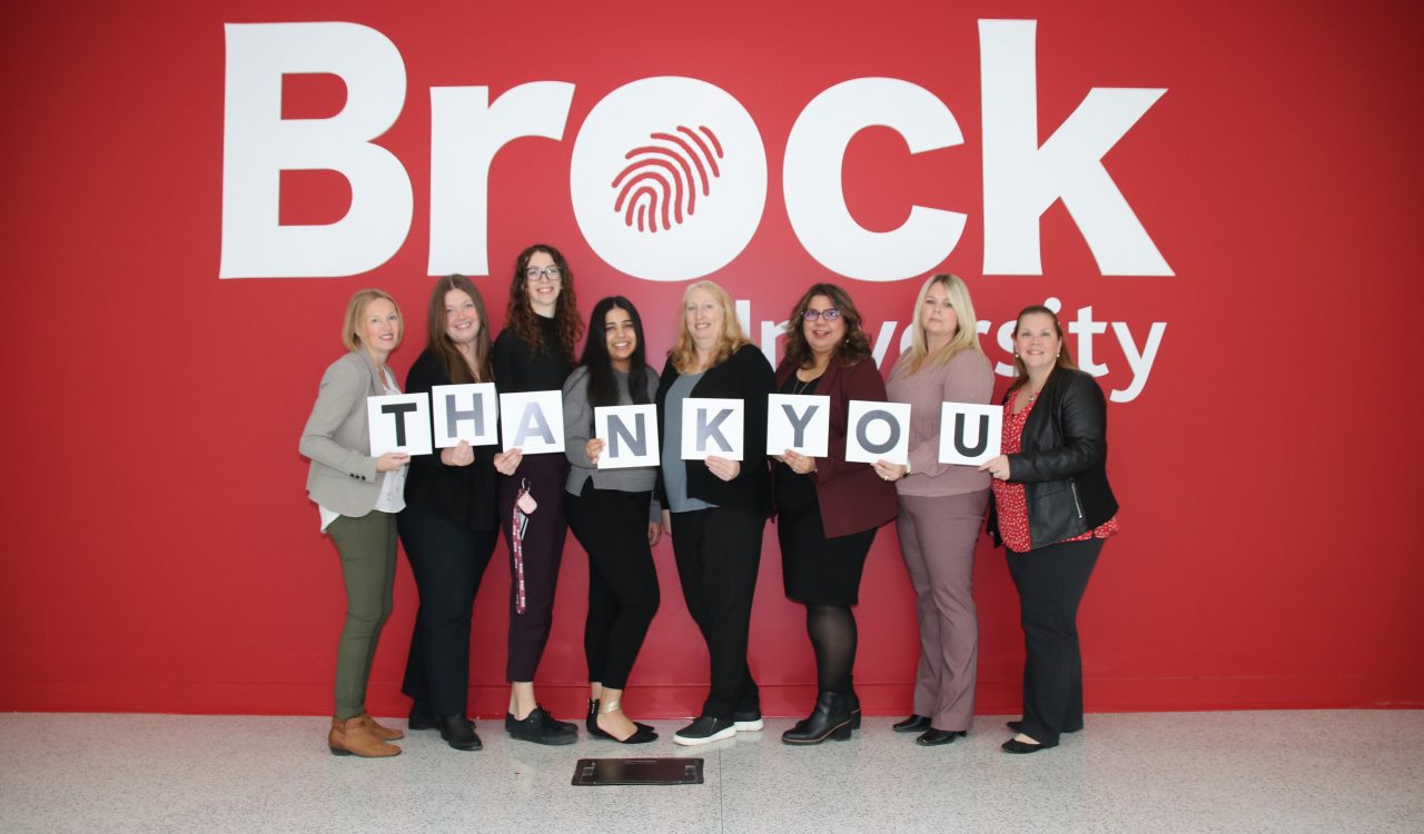 Brock’s Development and Donor Relations team holding a thank-you sign of individual letters in front of a Brock-branded wall.