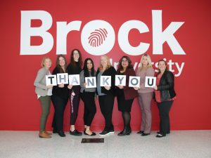 Brock’s Development and Donor Relations team holding a thank-you sign of individual letters in front of a Brock-branded wall.