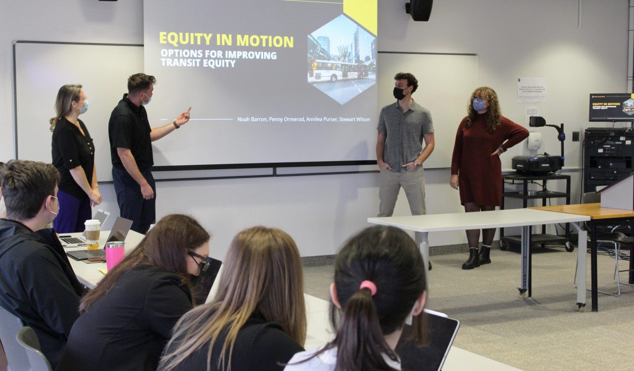 Four masked students stand in front a large projection screen with the title of their presentation, “Equity in Motion: Options for Improving Transit Equity.”