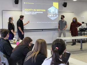Four masked students stand in front a large projection screen with the title of their presentation, “Equity in Motion: Options for Improving Transit Equity.”