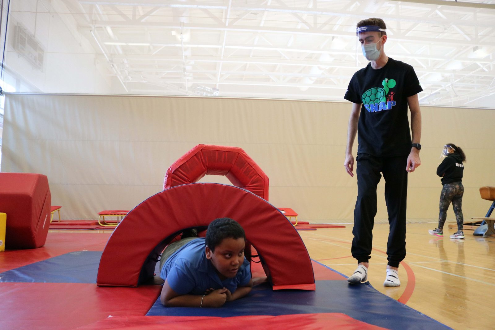 A child climbs through a padded archway under the supervision of a masked program co-ordinator in a gymnasium.