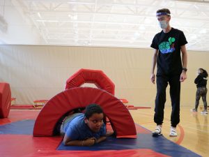 A child climbs through a padded archway under the supervision of a masked program co-ordinator in a gymnasium.