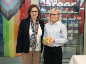 Two women stand together holding a small box wrapped in gold paper and bow.
