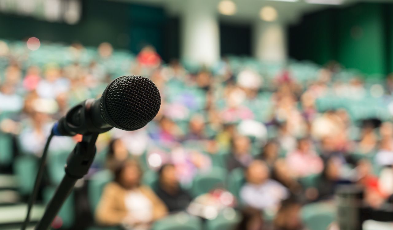 A focused shot of a microphone, with an audience in a lecture setting blurred in the background