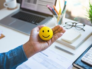 A yellow smiley face toy is held in someone’s palm. In the background is a desktop with a laptop, coffee mug, pad of paper, calculator and eyeglasses.