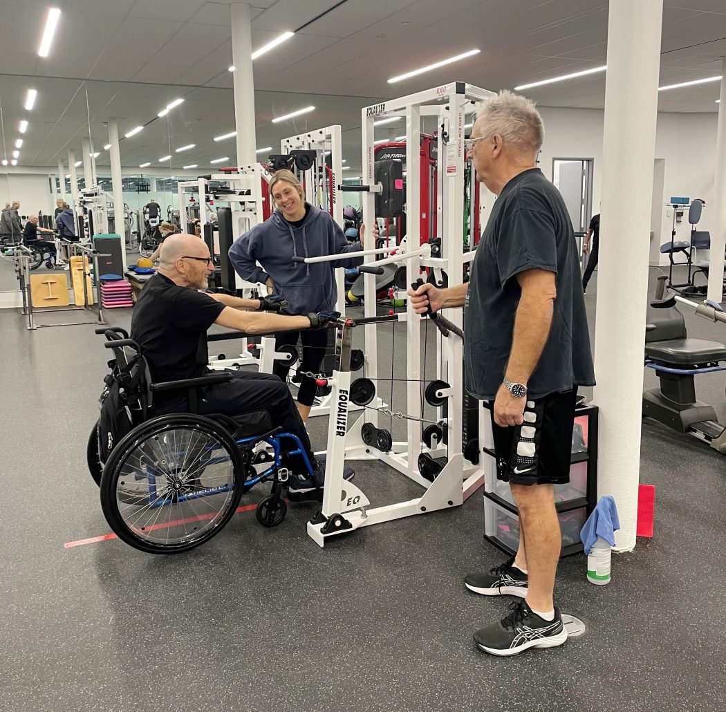 Bfit Centre Manager speaks with a Power Cord member exercising in a wheelchair while a SeniorFit member does standing exercises nearby.