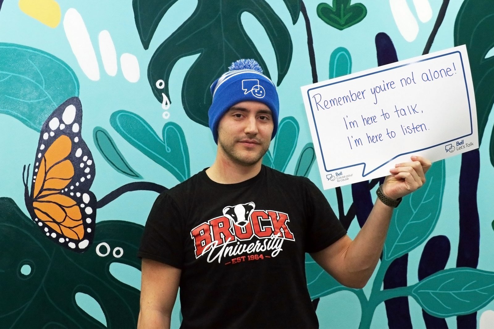 Brock University student Andrew Sheehan holds up a message of support he has written on a paper conversation bubble. He is wearing a Brock University T-shirt and Bell Let’s Talk Day toque. The message reads “Remember you’re not alone! I’m here to talk. I’m here to listen.”