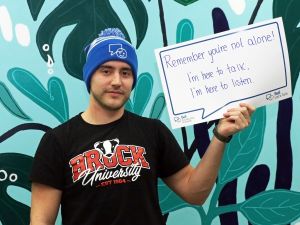 Brock University student Andrew Sheehan holds up a message of support he has written on a paper conversation bubble. He is wearing a Brock University T-shirt and Bell Let’s Talk Day toque. The message reads “Remember you’re not alone! I’m here to talk. I’m here to listen.”