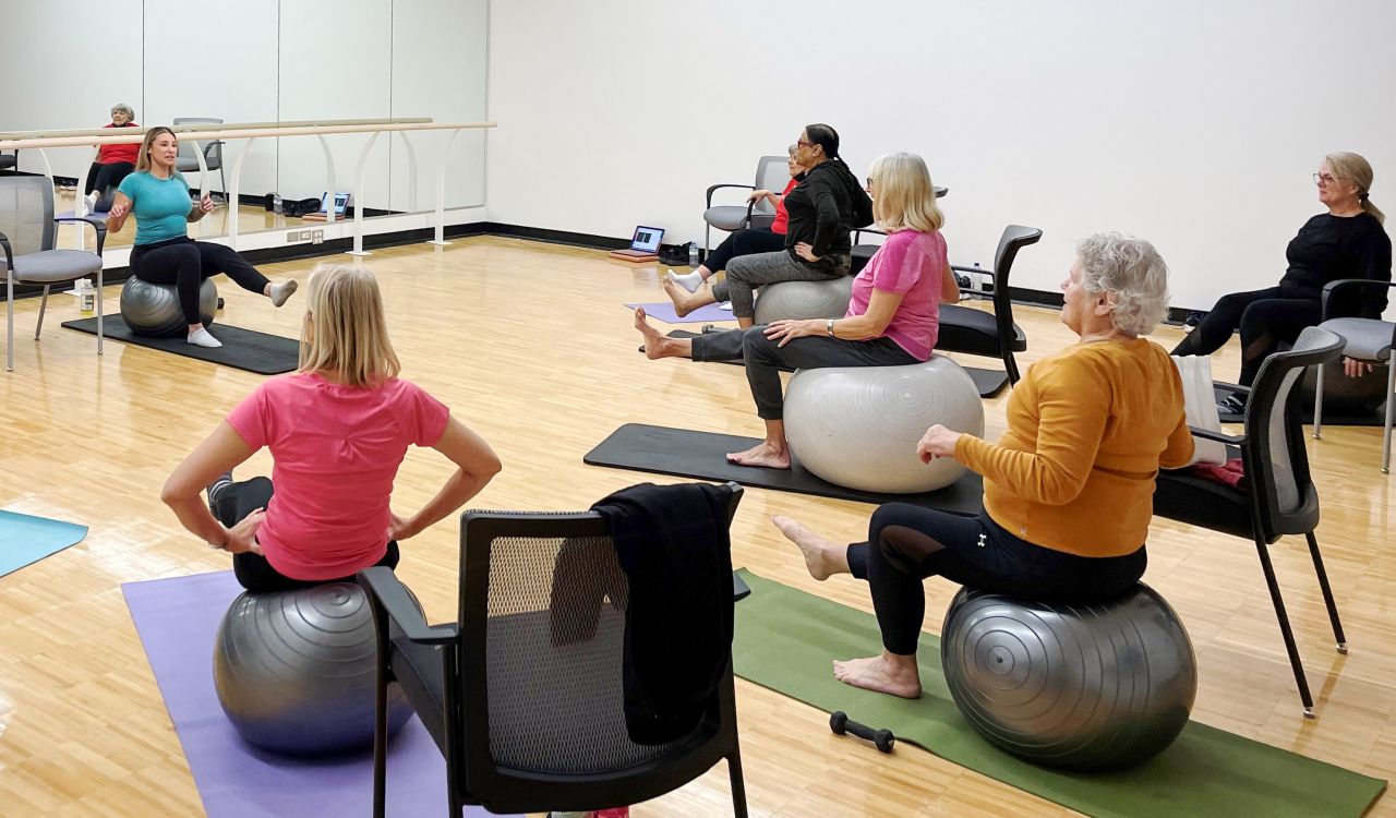 Bfit members participate in an exercise ball class led by a graduate student. Participants each sit on a yoga ball while watching the instructor.