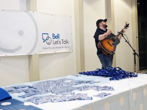 A student plays the guitar next to a table filled with Bell Let's Talk toques, pamphlets and lanyards.