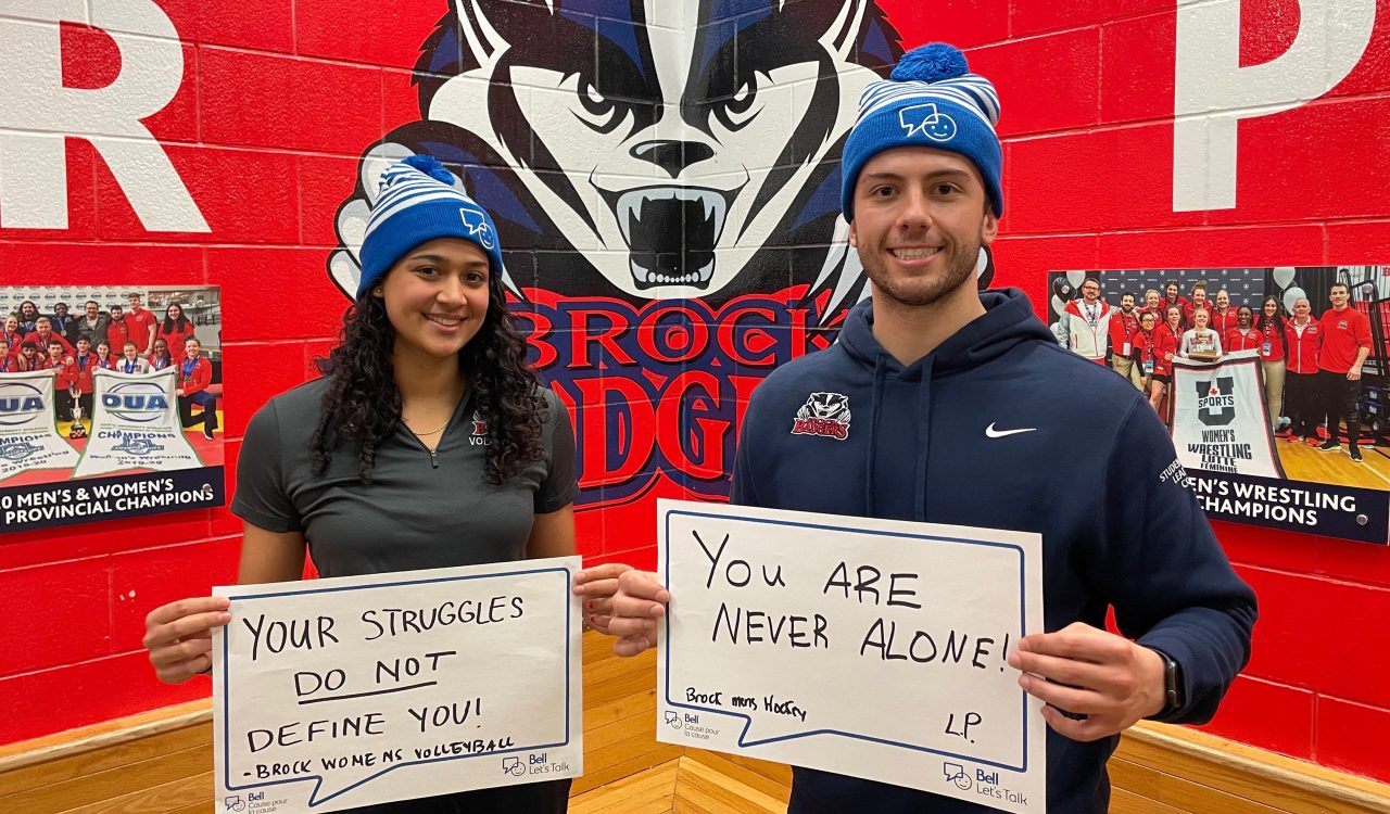 A woman and a man stand indoors holding signs with words of positive encouragement regarding mental health.