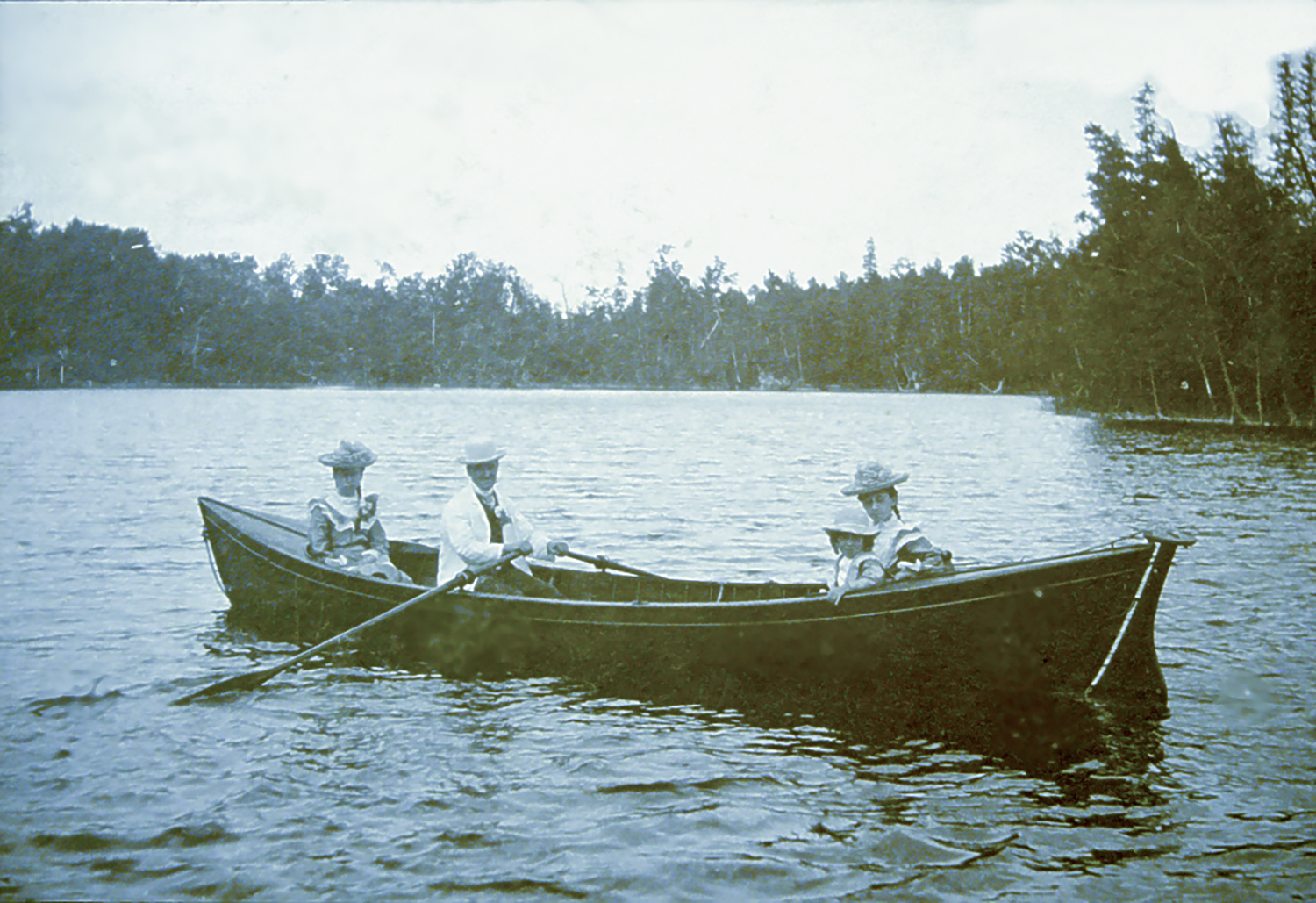 Three people in old-fashioned clothing sit in a canoe in the middle of a lake.