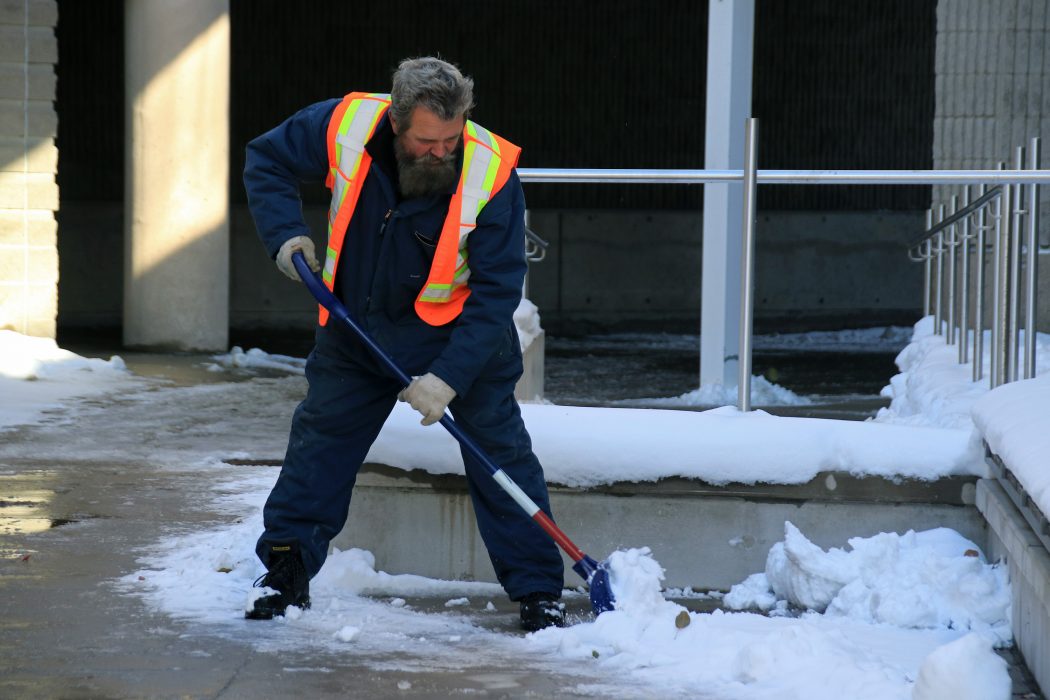 A man wearing an orange construction vest and blue work jumpsuit shovels show and ice from a sidewalk.