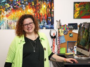 Diana deMan stands in her office at Brock University, with her hand resting on a raised standup desk. Behind her is a colourful painting of a tree-lined path.