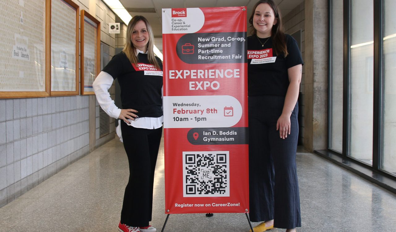 Two women in black t-shirts stand on either side of a large red sign in a hallway.