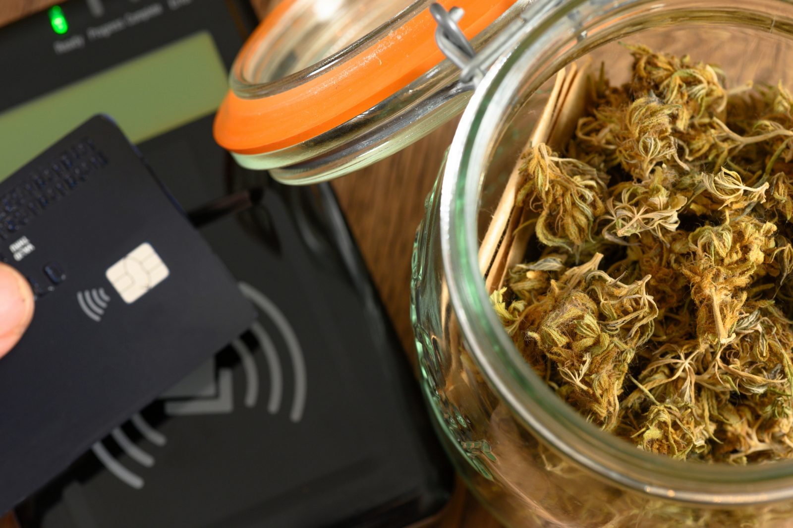 A hand taps a credit card over a contactless payment system beside a glass jar filled with cannabis.