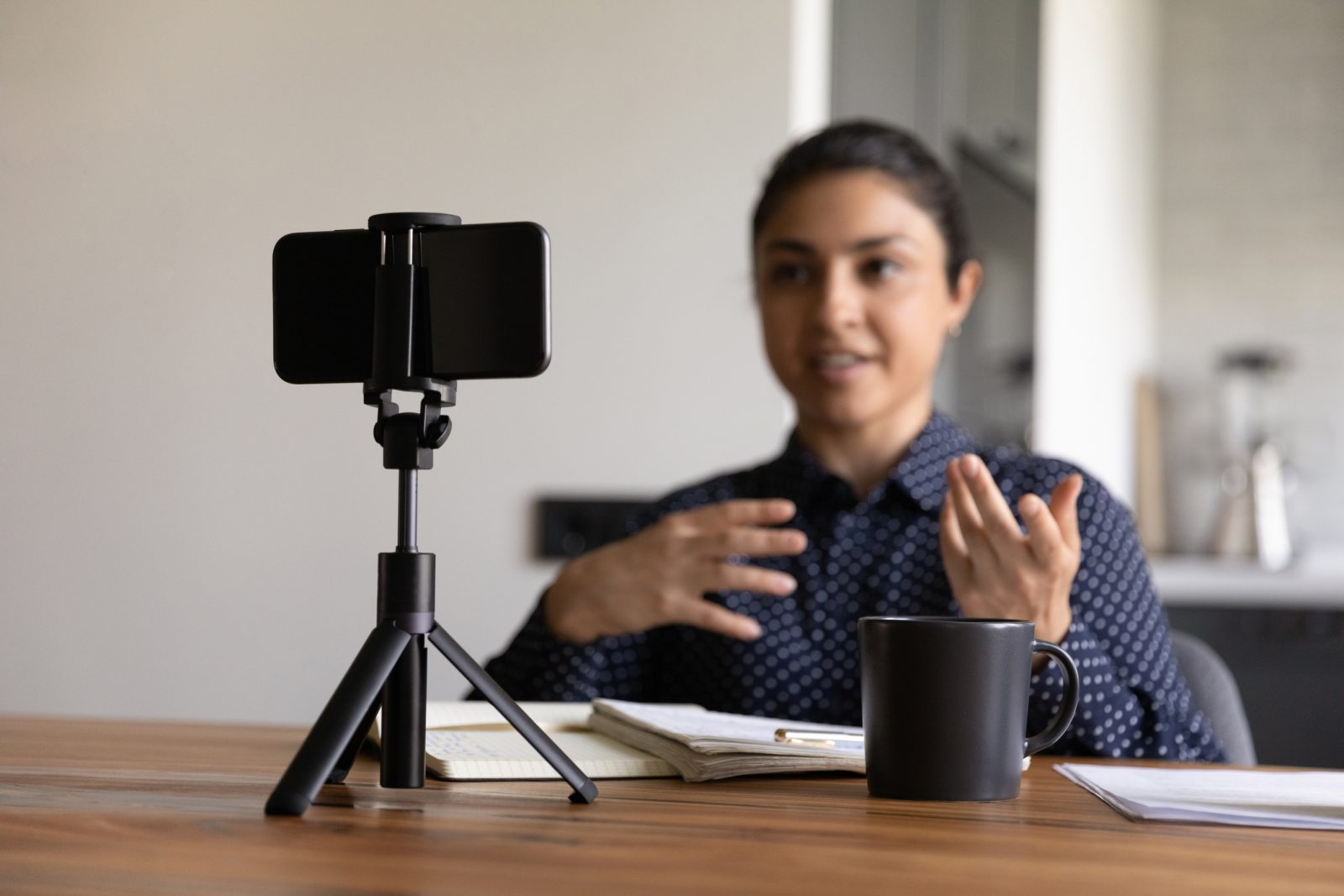 A woman sits at a table speaking at a phone on a tripod that is facing her.