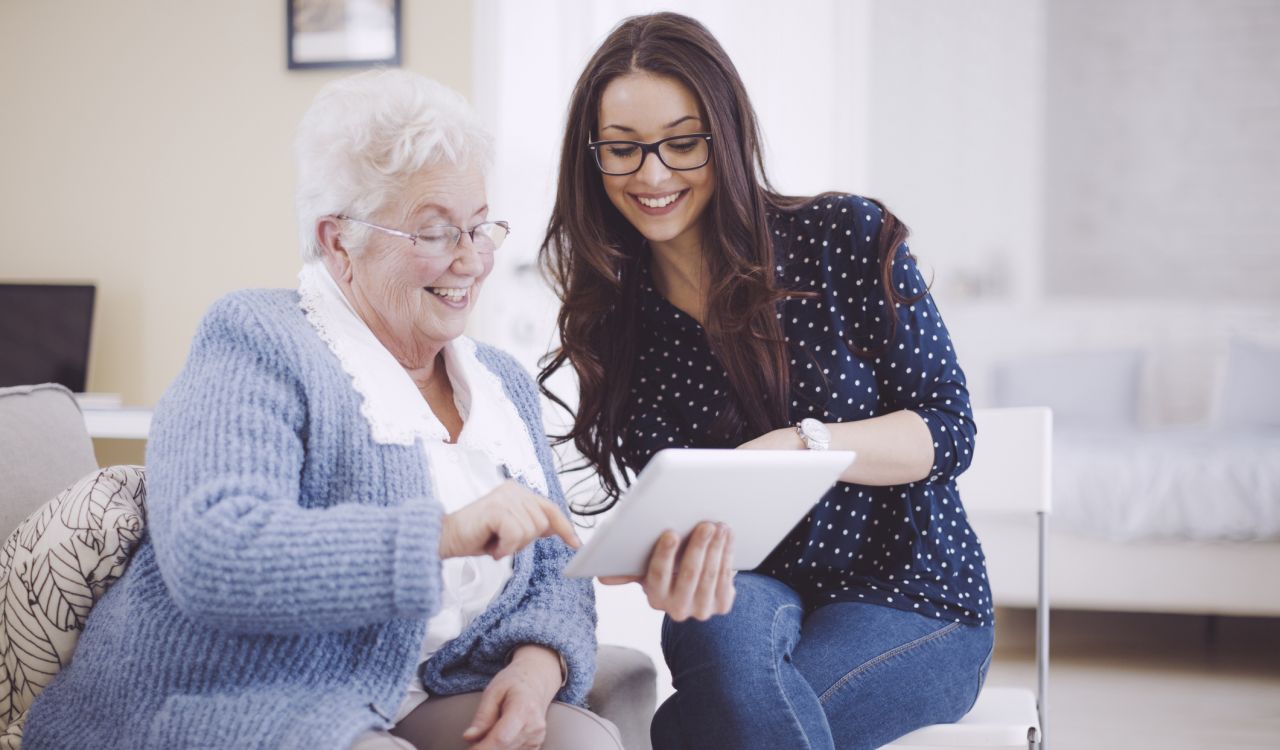 A smiling, seated older woman with short silver hair and glasses wearing a blue sweater points to a tablet being held by a smiling young women with long brown hair, glasses and wearing a blue pock dotted blouse sitting on her right in another chair also pointing at the tablet.