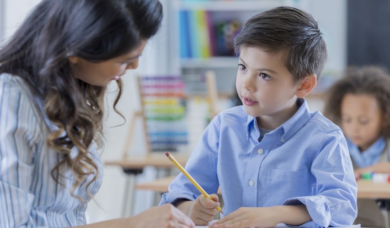 A young boy sits at a table with a pencil in hand and is taking instruction from a woman who is speaking to him while also seated.