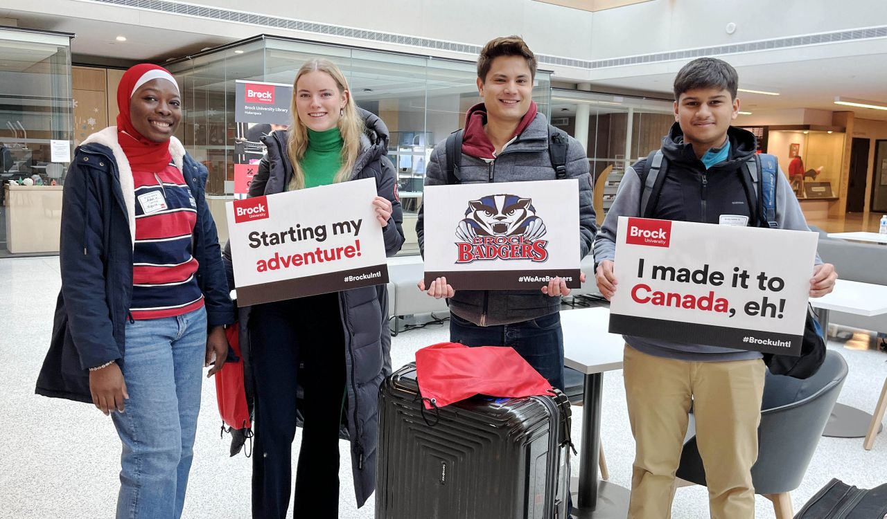 Four Brock University students stand behind suitcases holding cardboard signs in an indoor atrium.