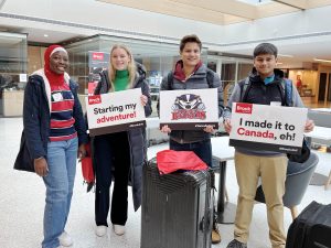 Four Brock University students stand behind suitcases holding cardboard signs in an indoor atrium.